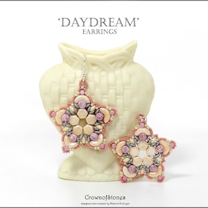 May include: Star-shaped earrings with a beaded design in shades of pink, cream, and silver. The earrings are labeled 'DAYDREAM' and are displayed in front of a cream-colored vase. The brand name 'CrownofStones' is also visible.