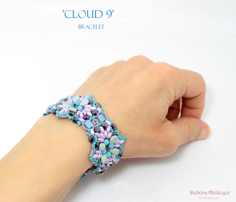 May include: A beaded bracelet with the text "CLOUD 9" and "BRACELET". The bracelet features a floral design with light blue, lavender, and dark purple beads. The bracelet is worn on a wrist.