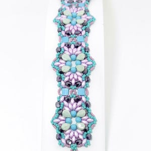 May include: A beaded bracelet with a floral design in shades of lavender, teal, and blue. The bracelet features the text "'CLOUD 9' BRACELET" at the top and the designer's name, Debora Hodoyer, at the bottom.