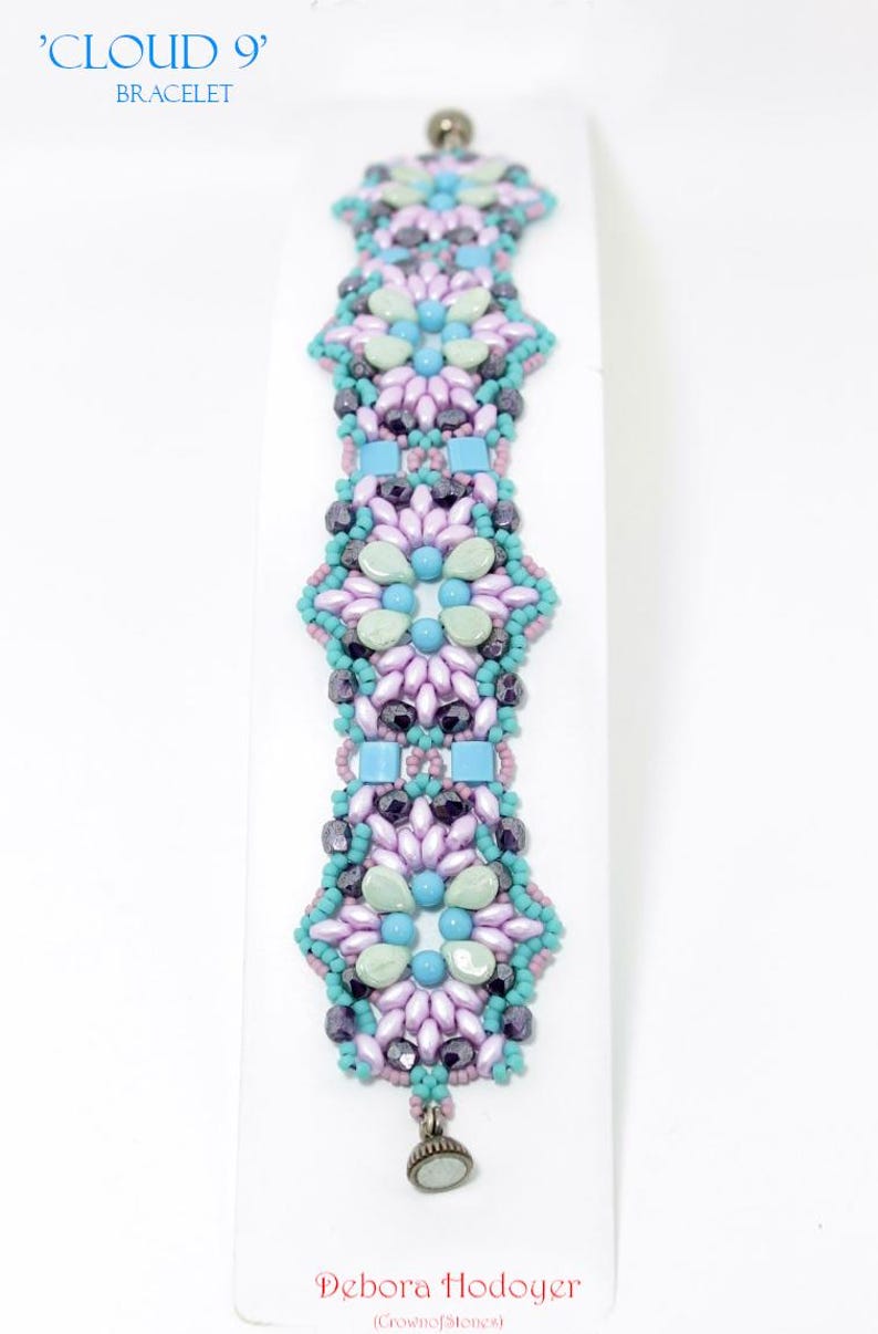 May include: A beaded bracelet with a floral design in shades of lavender, teal, and blue. The bracelet features the text "'CLOUD 9' BRACELET" at the top and the designer's name, Debora Hodoyer, at the bottom.