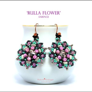 Könnte beinhalten: Florale Ohrringe mit dem Text "RULLA FLOWER" und "EARRINGS". Die Ohrringe haben ein Blumenmuster mit rosa, lila, türkis und silbernen Perlen. Die Ohrringe hängen an einer weißen Oberfläche.