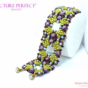Può includere: Un braccialetto di perline con perline verde oliva, viola e blu. Il braccialetto ha un design floreale ed è presentato su una superficie bianca. Le parole "PICTURE PERFECT" e "BRACELET" sono visibili.