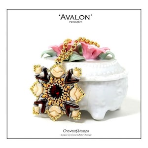 Könnte beinhalten: Eine goldfarbene Halskette mit einem aufwendigen Anhänger. Der Anhänger hat einen zentralen roten Stein, cremefarbene, braune und goldfarbene Details. Die Halskette wird auf einem weißen Keramikbehälter mit rosa Blumen präsentiert. Der Text auf dem Bild lautet 'AVALON' PENDANT und 'CrownofStones'.