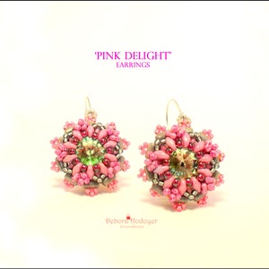 May include: Pink Delight earrings. These handmade earrings feature a floral design with pink, gray, and silver beads. The center of each earring has a green and iridescent crystal. The earrings are on a white background.