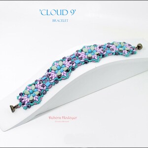 May include: A beaded bracelet with a floral design in shades of purple, teal, and blue. The bracelet is displayed on a white angled surface. The text "CLOUD 9" and "BRACELET" are in the upper left corner.