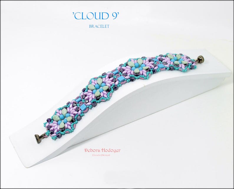 May include: A beaded bracelet with a floral design in shades of purple, teal, and blue. The bracelet is displayed on a white angled surface. The text "CLOUD 9" and "BRACELET" are in the upper left corner.