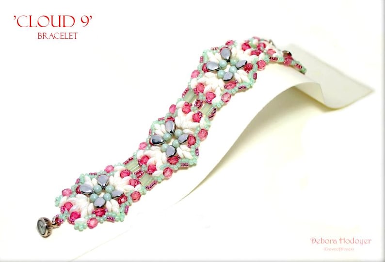 May include: A beaded bracelet with the text "CLOUD 9" and "BRACELET" in red. The bracelet features white, pink, and light blue beads. The bracelet is displayed on a white surface.