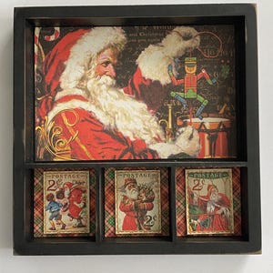 May include: A black framed Christmas-themed artwork featuring a vintage Santa Claus illustration. The artwork includes a large image of Santa and three postage stamp-style images of Santa. The stamps read "POSTAGE" and "2¢".