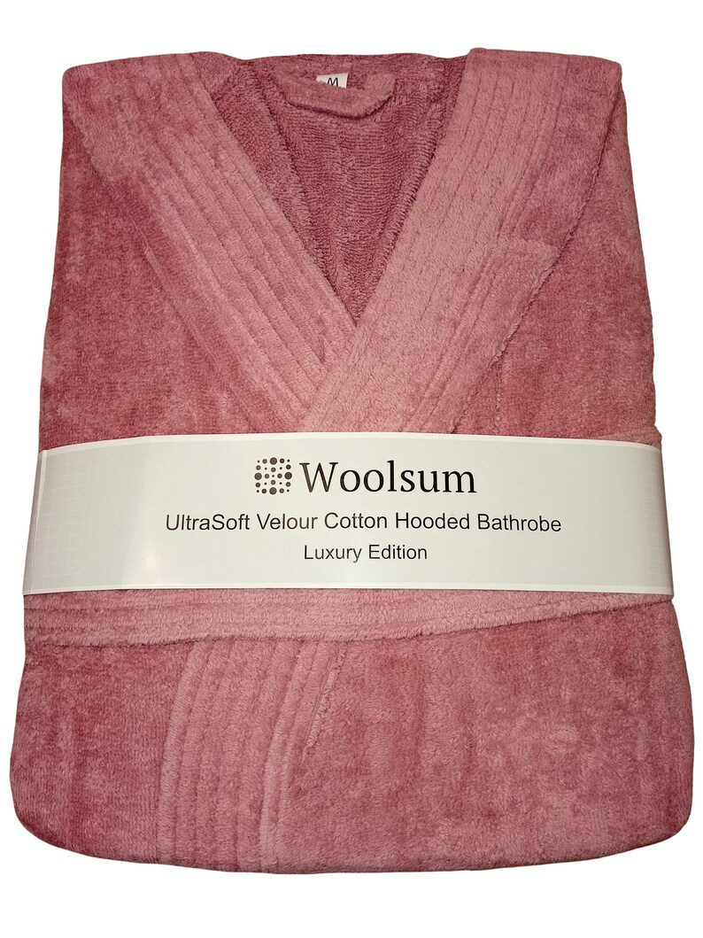 May include: A folded, dusty rose-colored hooded bathrobe. The bathrobe is wrapped with a white band that reads "Woolsium UltraSoft Velour Cotton Hooded Bathrobe Luxury Edition."