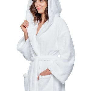 May include: A white hooded bathrobe with a matching belt and pockets. The plush robe is made of a soft, absorbent material, perfect for after a shower or a relaxing day at home. The model is wearing the robe.