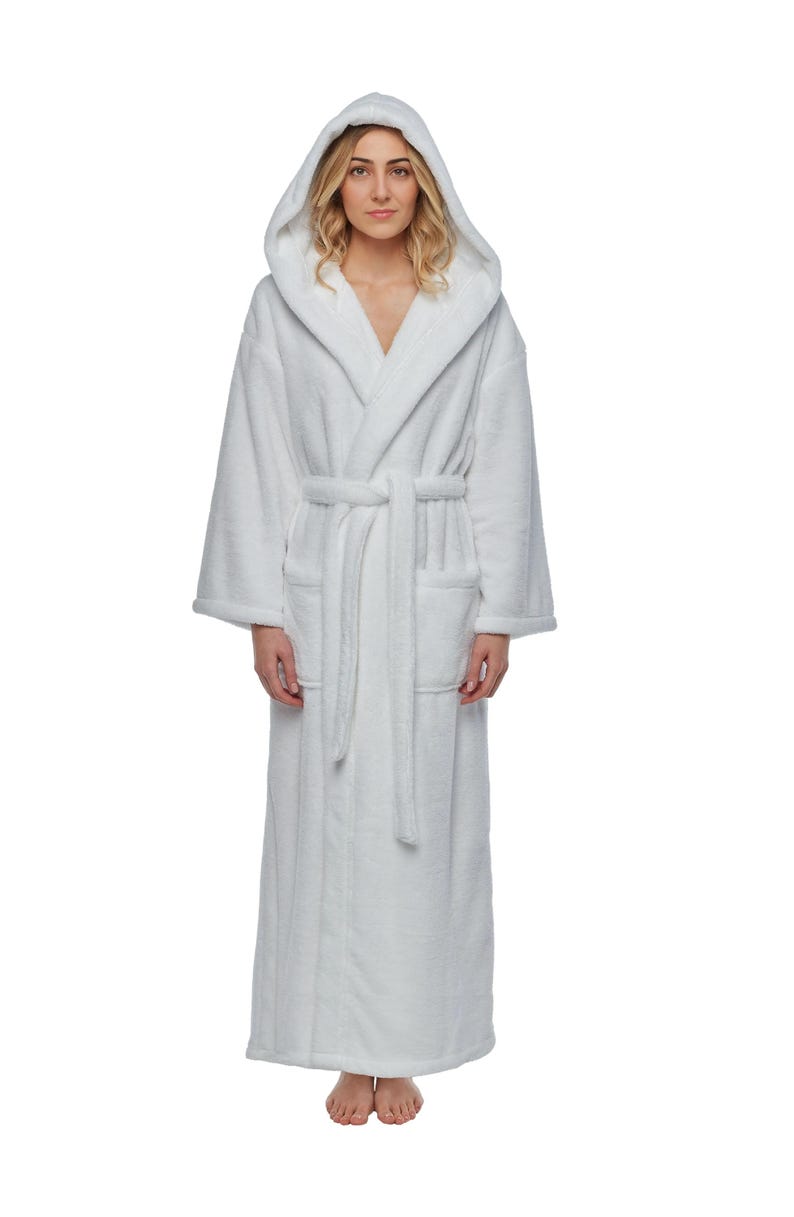 May include: A full-length white hooded bathrobe with a matching belt and pockets. The robe is made of a soft, plush material. The model is wearing the robe.