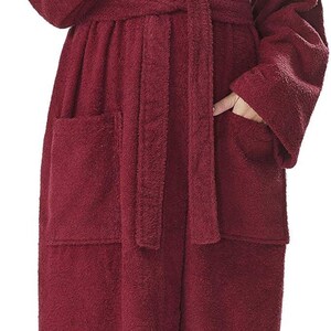 May include: A full-length, hooded burgundy terry cloth robe with a matching belt and pockets. The robe is worn by a person with blonde hair. The robe is made of a soft, absorbent material, perfect for after a shower or bath.