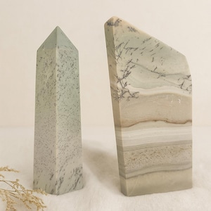 May include: Two stone obelisks. One is a tall, rectangular shape with a pointed top and a speckled, light green color. The other is a taller, angled shape with layered beige, white, and brown tones, and a dark, tree-like pattern.