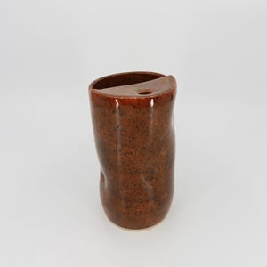May include: A cylindrical ceramic vase with a glossy, speckled brown glaze. The vessel features an irregular, organic shape with a flat, semi-circular top section that includes a single small circular opening alongside a larger open cavity.