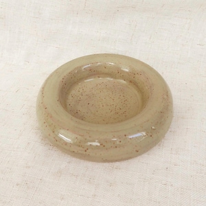 Op de afbeelding: Een keramische asbak met een cirkelvormig ontwerp. De asbak is lichtbeige van kleur met kleine bruine spikkels. De asbak heeft een glanzende afwerking en een verzonken midden.