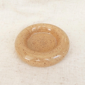 May include: A small, round ceramic ashtray with a light tan glaze and speckled brown details. The ashtray has a raised outer rim and a recessed centre. The design is simple and functional.