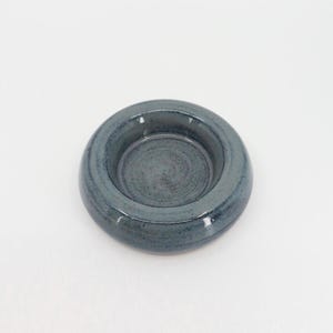 Pode incluir: Um pequeno prato circular de cerâmica com uma borda espessa e arredondada e um centro rebaixado. A peça apresenta um esmalte brilhante e manchado em tons de azul ardósia profundo e cinza carvão, com variações sutis de cor e textura.