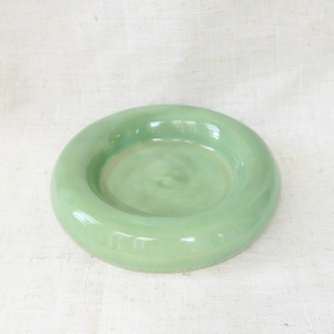 May include: A circular, pale green ceramic dish with a raised rim and a shallow central area. The dish has a glossy finish and is placed on a white, textured surface. The shape is a complete circle.