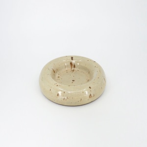 Può includere: Un piatto in ceramica circolare con una finitura liscia e lucida in tonalità beige chiaro. La superficie presenta macchie marroni sparse, con un centro leggermente incavato e un bordo spesso e arrotondato che crea un aspetto a ciambella.
