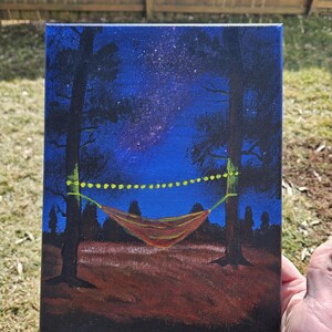 May include: A painting of a nighttime scene with a hammock strung between two trees. The sky is a deep blue with visible stars. A string of yellow lights hangs above the hammock, which is a reddish-orange color. The ground is a dark brown.