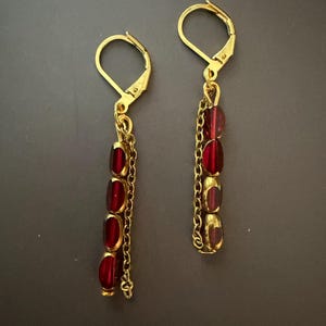 May include: A pair of gold-colored dangle earrings. Each earring features a lever-back closure, a chain, and four oval, deep red beads. The earrings are displayed against a dark gray background.