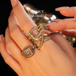 Puede incluir: Una mano con varios anillos dorados. Un anillo presenta una piedra rosa cuadrada con un diseño de calavera. Otros anillos incluyen un diseño articulado con pequeñas piedras transparentes y una cadena de cuentas. El texto "YUMMY ShopStudio" es visible.