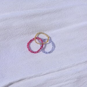 以下が含まれることがあります： ピンク、ゴールド、ライトブルーのビーズリング3つが、白いテクスチャのある表面に配置されています。リングは小さく光沢のあるビーズでできており、その円形が背景に対して明確に定義されています。