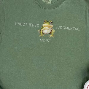 Op de afbeelding: Olijfgroen T-shirt met een kikker-afbeelding en de tekst "Unbothered, Judgmental, Moist". De afbeelding bevat lichtblauwe denim jeans en witte canvas sneakers met rode accenten. Een kleine groene plant is ook zichtbaar.