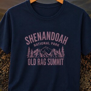 May include: A navy blue short-sleeve t-shirt featuring a light purple graphic print. The design includes the text SHENANDOAH NATIONAL PARK and OLD RAG SUMMIT above and below a stylized illustration of mountain peaks and evergreen trees.