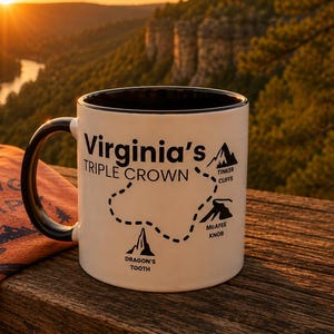 May include: A white ceramic mug with a black handle and interior, featuring the text Virginia's Triple Crown, a dotted trail map, and illustrations of Tinker Cliffs, McAfee Knob, and Dragon's Tooth, alongside a rust-colored shirt with hiking text.