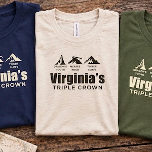 May include: Three crew neck t-shirts in navy blue, cream, and olive green. Each shirt features a graphic of three mountain peaks labeled Dragon's Tooth, McAfee Knob, and Tinker Cliffs, with the text Virginia's Triple Crown printed below.