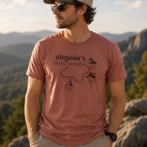 Peut inclure: Un t-shirt couleur rouille avec le texte "Virginia's Triple Crown" et une carte de trois sommets montagneux. Le t-shirt est associé à un short kaki, une casquette de camionneur verte et beige et des lunettes de soleil. L'arrière-plan présente une chaîne de montagnes.