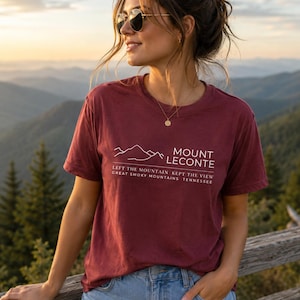 Może przedstawiać: Bordowa koszulka z białym napisem i grafiką gór. Tekst brzmi "Mount LeConte, Left the Mountain Kept the View, Great Smoky Mountains Tennessee." Koszulka jest połączona z dżinsowymi szortami i okularami przeciwsłonecznymi.