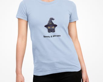Op de afbeelding: Lichtblauw T-shirt met een cartoon zwarte kat met een heksenhoed. De kat heeft gele ogen en zit. De tekst "Spooky & Adorable" staat eronder. Het shirt heeft korte mouwen.