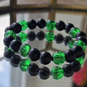 May include: A bracelet featuring alternating black and green beads. The beads are round and appear to be made of glass or a similar material. The bracelet is displayed on a reflective surface, creating a mirrored effect.