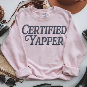 Könnte beinhalten: Ein hellrosa Sweatshirt mit dem Text "CERTIFIED YAPPER" in dunkelgrauer Schrift. Das langärmlige Sweatshirt mit Rundhalsausschnitt ist an den Bündchen umgeschlagen. Das Sweatshirt liegt auf einer weißen Oberfläche, mit Accessoires im Hintergrund.