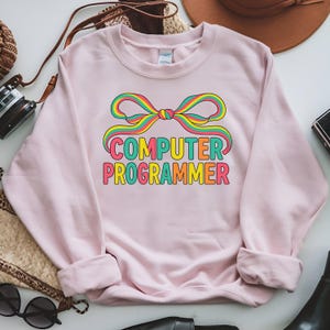 Op de afbeelding: Een lichtroze sweatshirt met de tekst "COMPUTER PROGRAMMER" in kleurrijke blokletters. Boven de tekst staat een regenboogstrikontwerp. De sweatshirt heeft lange mouwen en een ronde hals.