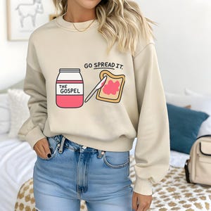 May include: Cream-coloured sweatshirt featuring a graphic of a jar labelled "THE GOSPEL", a slice of toast with pink spread, and the text "GO SPREAD IT." The sweatshirt is worn with blue jeans.