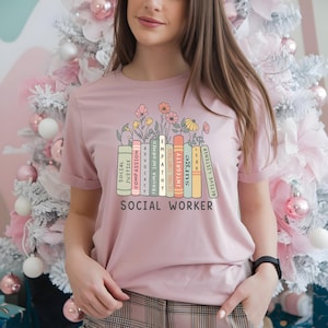 Pode incluir: Camiseta rosa claro com um desenho de livros coloridos e flores. Os livros têm palavras como "Empatia" e "Integridade". O texto "Social Worker" está abaixo.