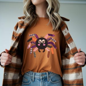Op de afbeelding: Een oranje t-shirt met een patchwork spinnenontwerp met paarse, oranje en zwarte accenten. De spin heeft grote ogen en een lachend gezicht. De persoon draagt een bruin en tan geruit jack en een blauwe jeans.
