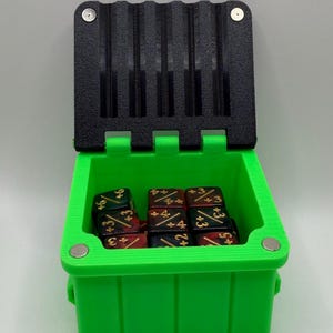 May include: A bright green dice box with a black lid, open to reveal several dice. The dice are black with gold markings and red accents. The box has a rectangular shape with a hinged lid and magnetic closures.