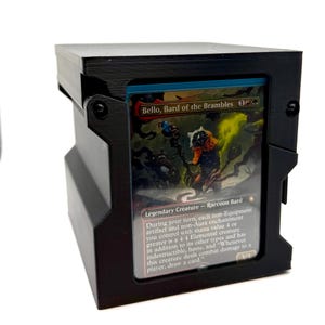 May include: Black card holder with a hinged lid, holding a Magic: The Gathering card. The card features the text "Bello, Bard of the Brambles" and a raccoon bard illustration. The card holder is made of black material.