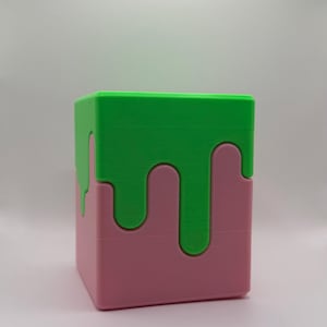 May include: A 3D-printed cube puzzle in pink and green. The top and sections of the sides are green, with a dripping effect over the pink base. The puzzle is a fun, colourful toy.