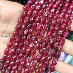 May include: A collection of oval-shaped, deep red gemstone beads. The beads are strung together, ready for use in jewellery making. The polished beads have a glossy finish. The image shows multiple strands, highlighting the colour and shape of the gemstones.