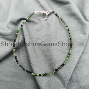 May include: A beaded necklace featuring small, faceted, multicolored gemstone beads in shades of green, black, and deep pink. The necklace is secured with a silver-toned metal lobster claw clasp and a short extender chain at the top center.