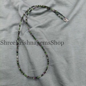 May include: A beaded necklace with a mix of green, black, white, and red beads. The necklace is laid on a gray surface. The text "ShreeKrishnagemsShop" is printed on the surface.