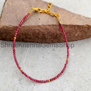 Könnte beinhalten: Ein zartes Armband mit kleinen, facettierten dunkelroten Perlen in kreisförmiger Anordnung, akzentuiert durch winzige goldfarbene Metallperlen in Abständen. Das Schmuckstück verfügt über eine goldfarbene Metallverlängerungskette mit Karabinerverschluss.