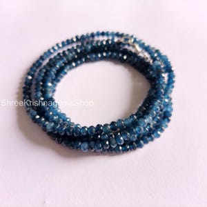 Puede incluir: Una colección de cuentas de piedras preciosas azul oscuro facetadas, ensartadas para formar un collar o una pulsera. Las pequeñas cuentas, de tamaño uniforme, crean una apariencia delicada y elegante. La joya se exhibe sobre una superficie blanca.