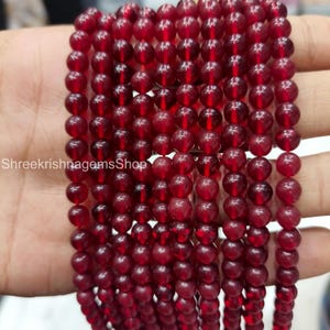Puede incluir: Hebras de cuentas translúcidas de color rojo intenso. Las cuentas son redondas y parecen estar ensartadas. La imagen está bien iluminada, resaltando el color y la textura de las cuentas. El texto "ShreekrishnagemsShop" es visible en la esquina inferior izquierda.