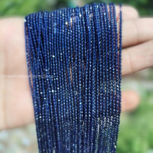 Op de afbeelding: Close-up van een bundel donkerblauwe edelsteen kralen. De kleine, gefacetteerde kralen zijn aan elkaar geregen, waardoor een glinsterend effect ontstaat. De afbeelding is goed belicht, waardoor de kleur en textuur van de kralen worden benadrukt. De tekst "ShreeKrishnaGemsShop" is zichtbaar.